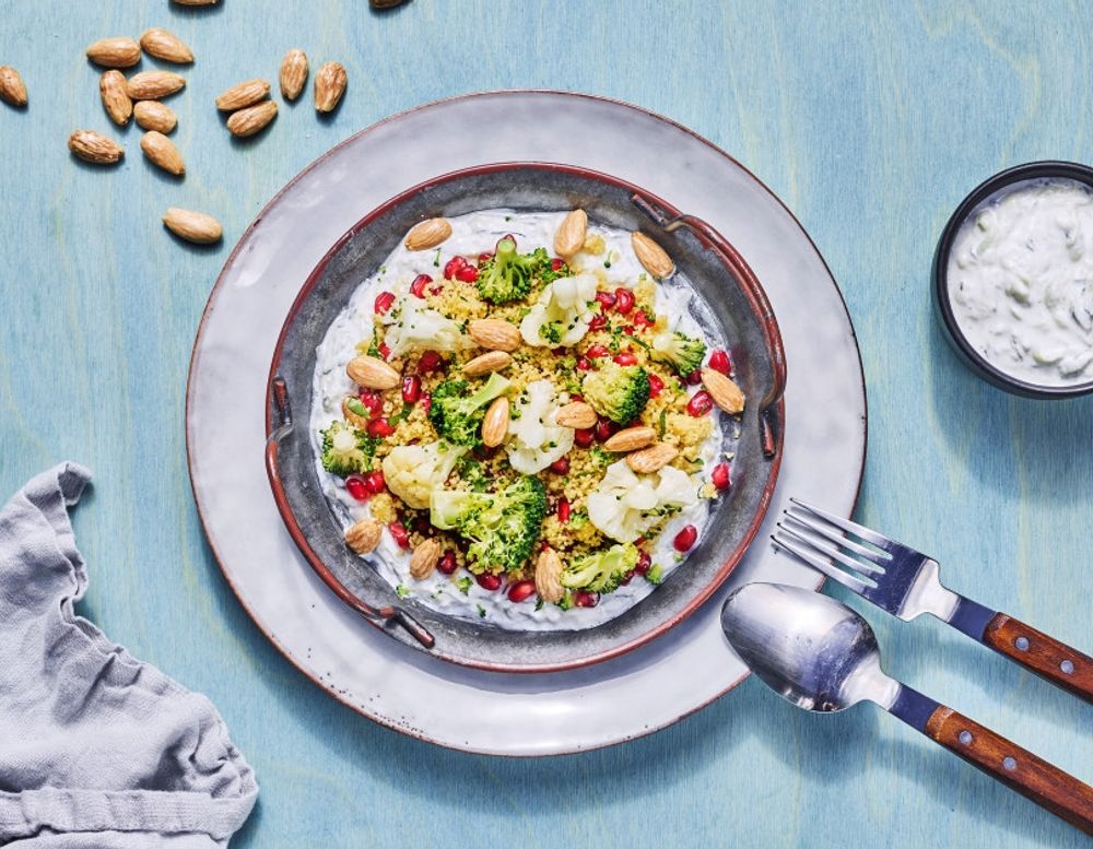 Salat med blomkål, brokkoli, quinoa, granateplekjerner og ristede mandler servert på en seng av yoghurt, med en skål urtedressing ved siden av på et lyseblått bord.
