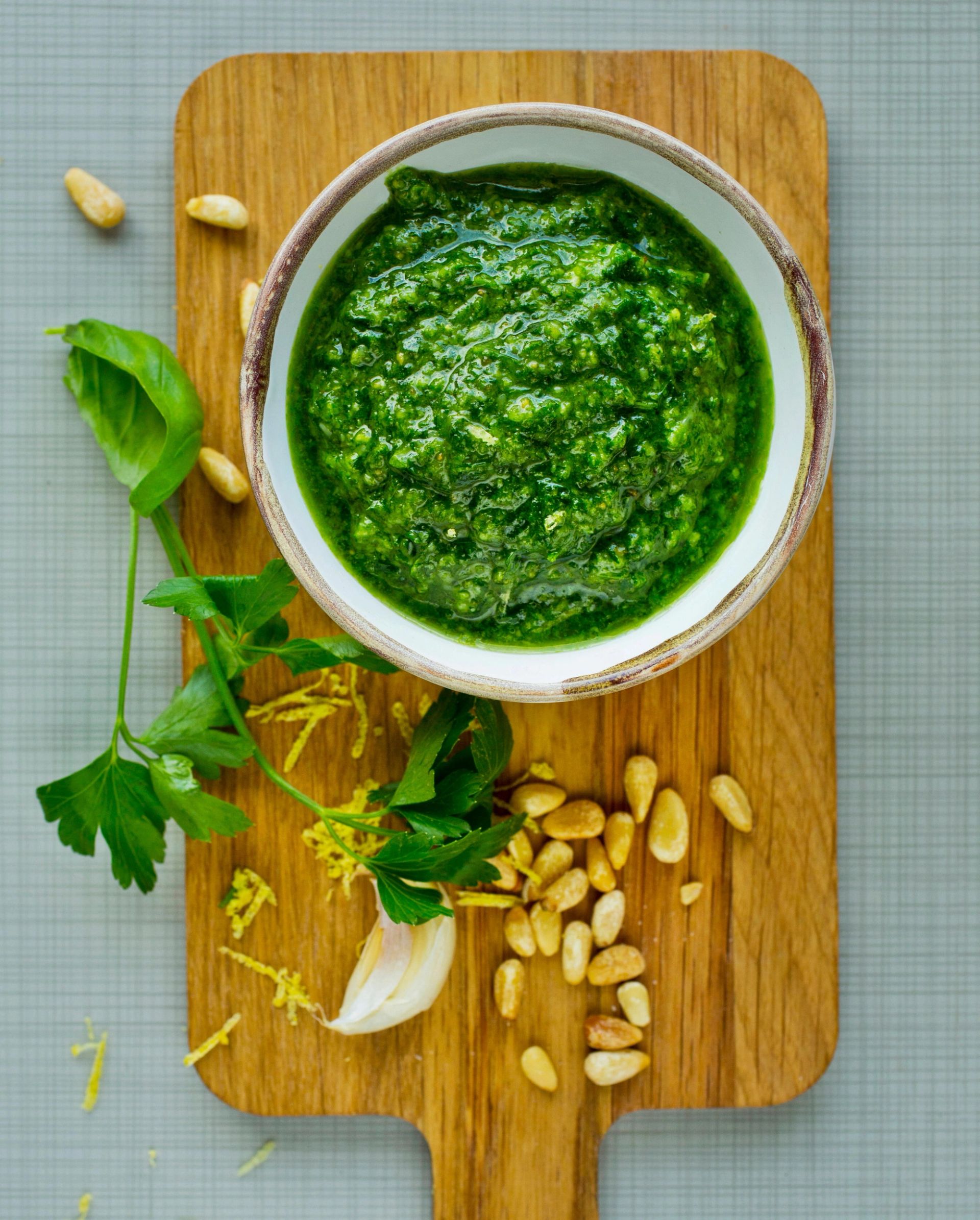 En bolle med frisk, grønn persillepesto står på et trefat. Rundt bollen ligger persilleblader, pinjekjerner, hvitløksfedd og litt finrevet sitronskall som ingredienser til pestoen.