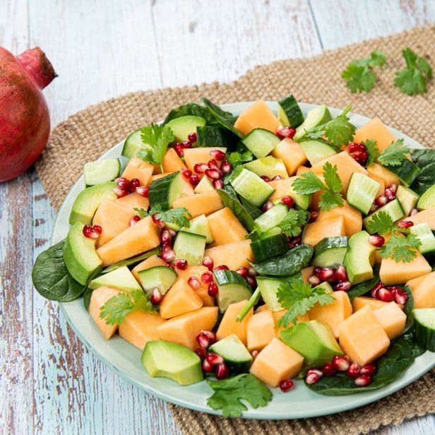 Salat med spinatblader toppet med avokado, terninger av cantaloupemelon, agurk, granateplekjerner og fersk koriander, servert på en lys tallerken på rustikk bordflate.