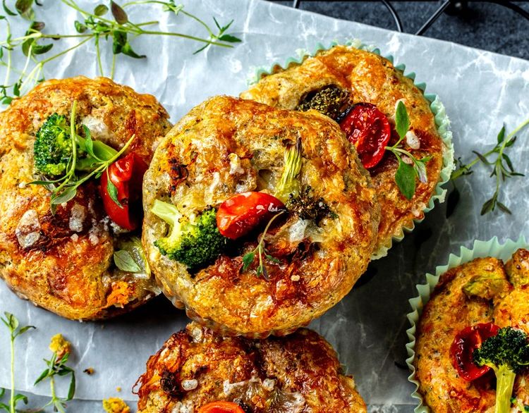 Salte grønnsaksmuffins med brokkoli, cherrytomater og urter, servert i papirformer på bakepapir, med fersk timian og oregano rundt.