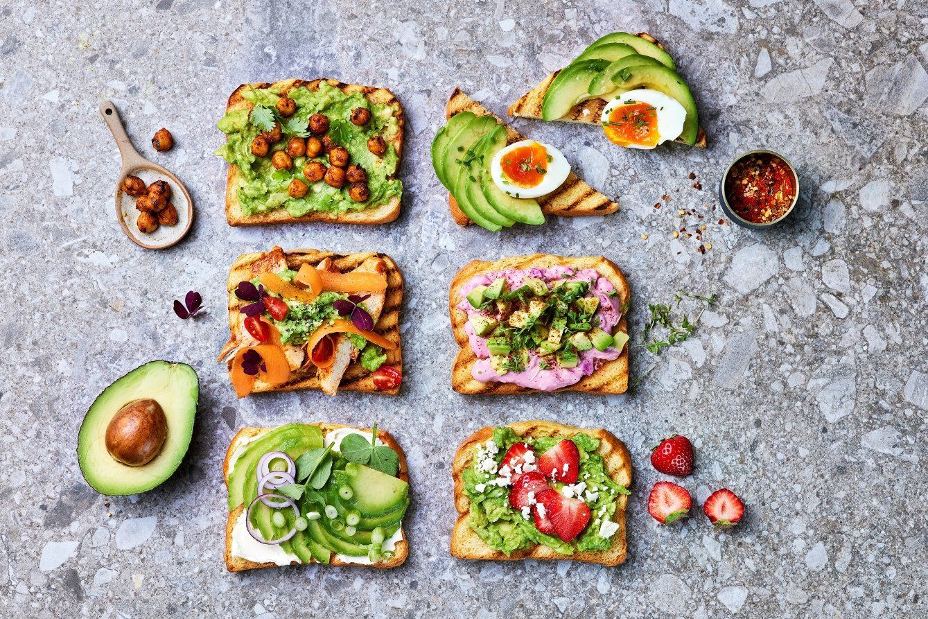 Seks forskjellige toast med avokado toppet med blant annet kikerter, egg, rødbethummus, grønnsaker, cherrytomater og jordbær. Servert på en steingrå overflate med fersk avokado og små tilbehørsskåler rundt.