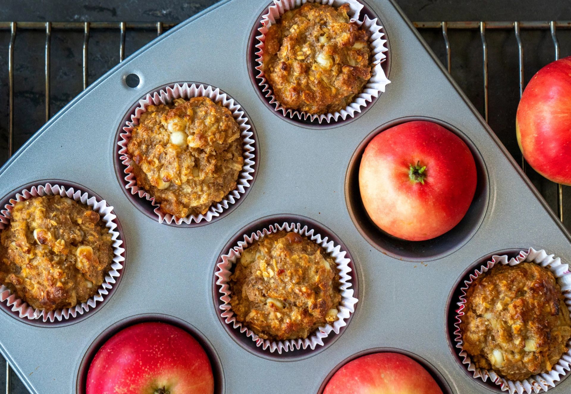 Grove eplemuffins i papirformer i en muffinsform på et bakerist, med friske røde epler rundt, avkjølt på et mørkt bord.
