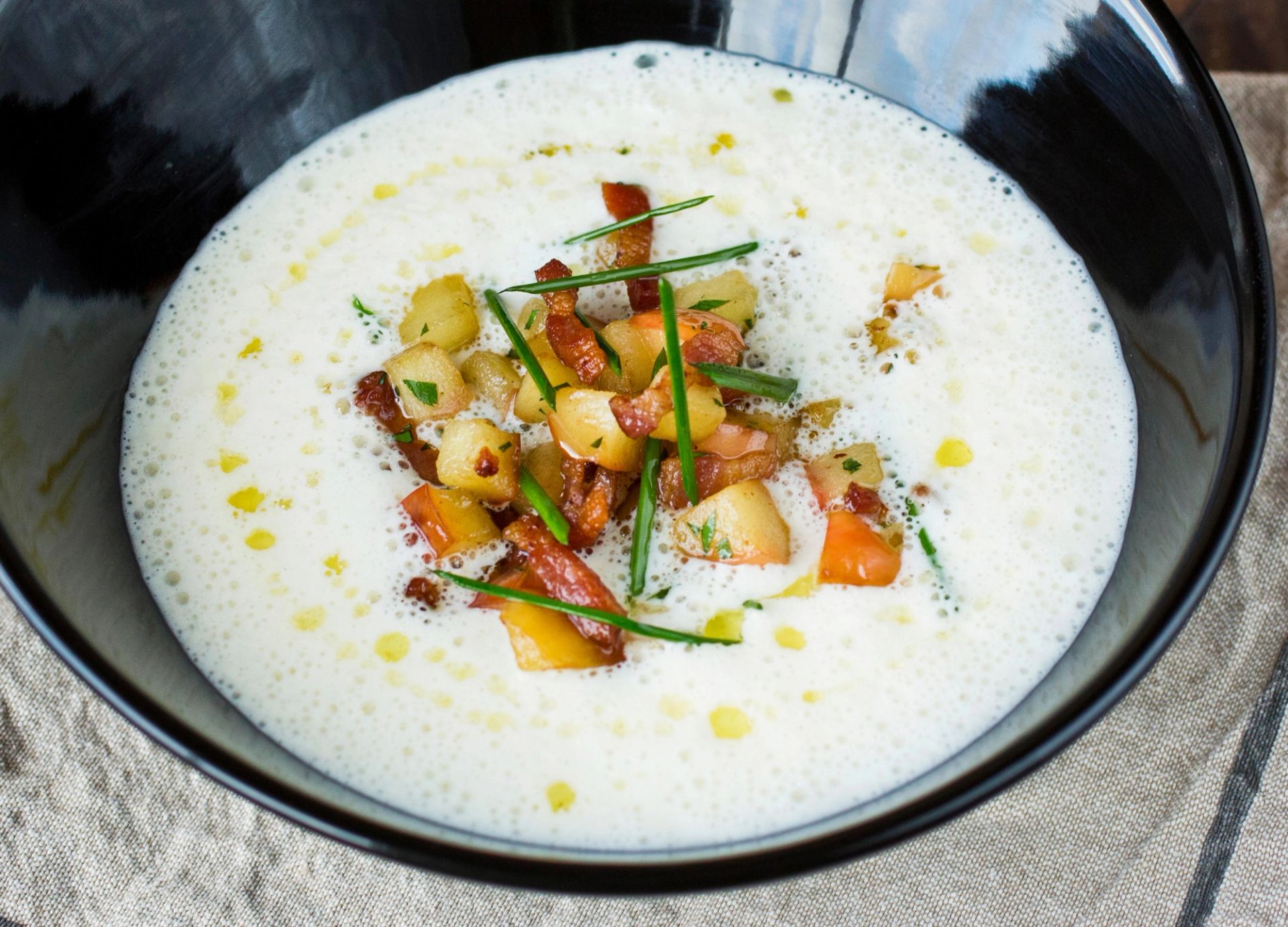 En dyp grå skål fylt med kremet, lys sellerirotsuppe. Midt i suppen ligger en topping av sprøstekte epler, små biter av stekt bacon eller røkt kjøtt, samt tynne strimler av gressløk. Det ligger små dråper med olje på overflaten av suppen, og skålen står på en lys tekstilduk.