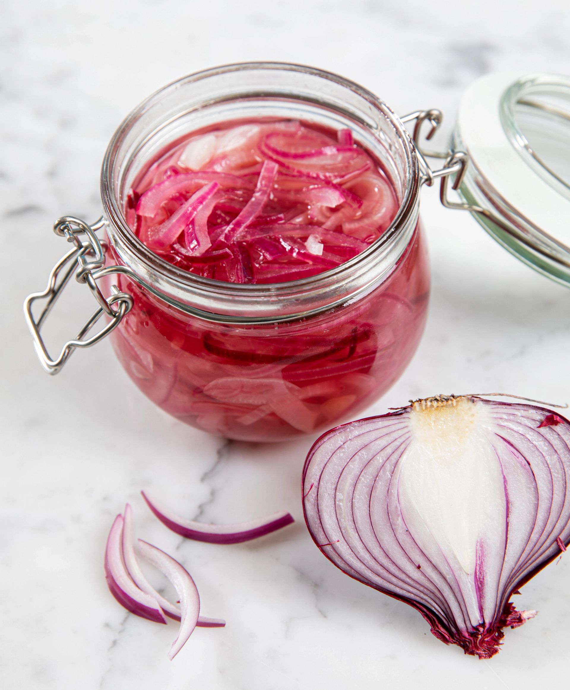 Et glass med syltet rødløk fylt med tynne lilla løkringer i rosa lake, med en halv rødløk og noen løkskiver liggende ved siden av på en lys marmorbakgrunn.