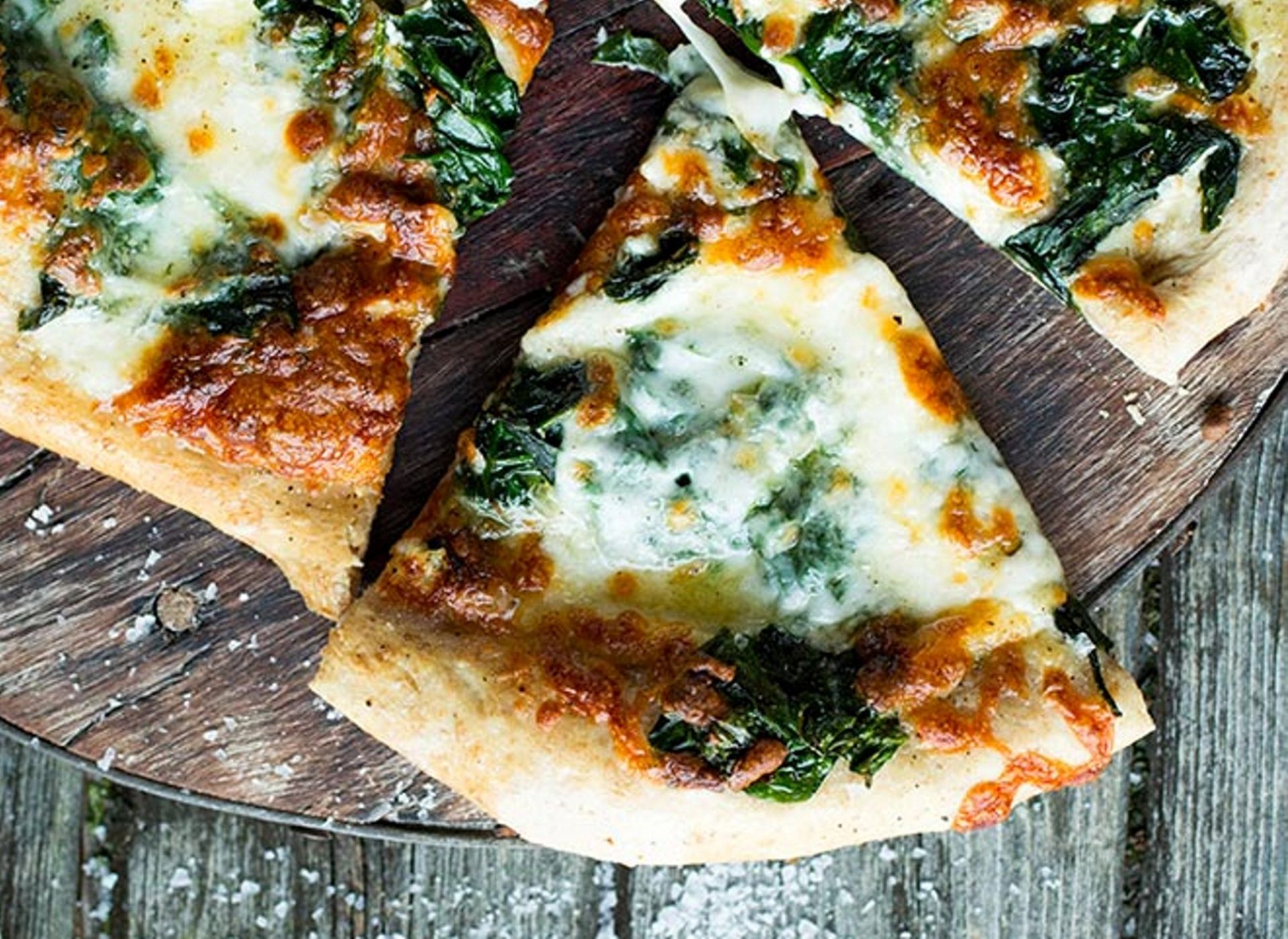 Et nærbilde av tre pizzastykker med hvit saus (pizza bianca) plassert på en rund trefjøl. Pizzaen er toppet med smeltet, gyllenbrent mozzarella og rikelig med grønn spinat.