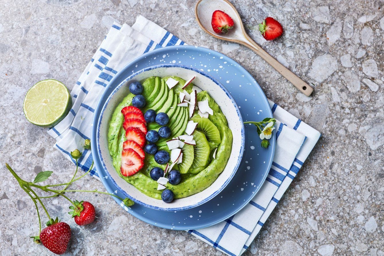 Kremet grønn smoothie laget av avokado, toppet med kiwi, jordbær, blåbær og kokosflak. Servert i en hvit bolle på blått fat med friske jordbær og lime ved siden av.