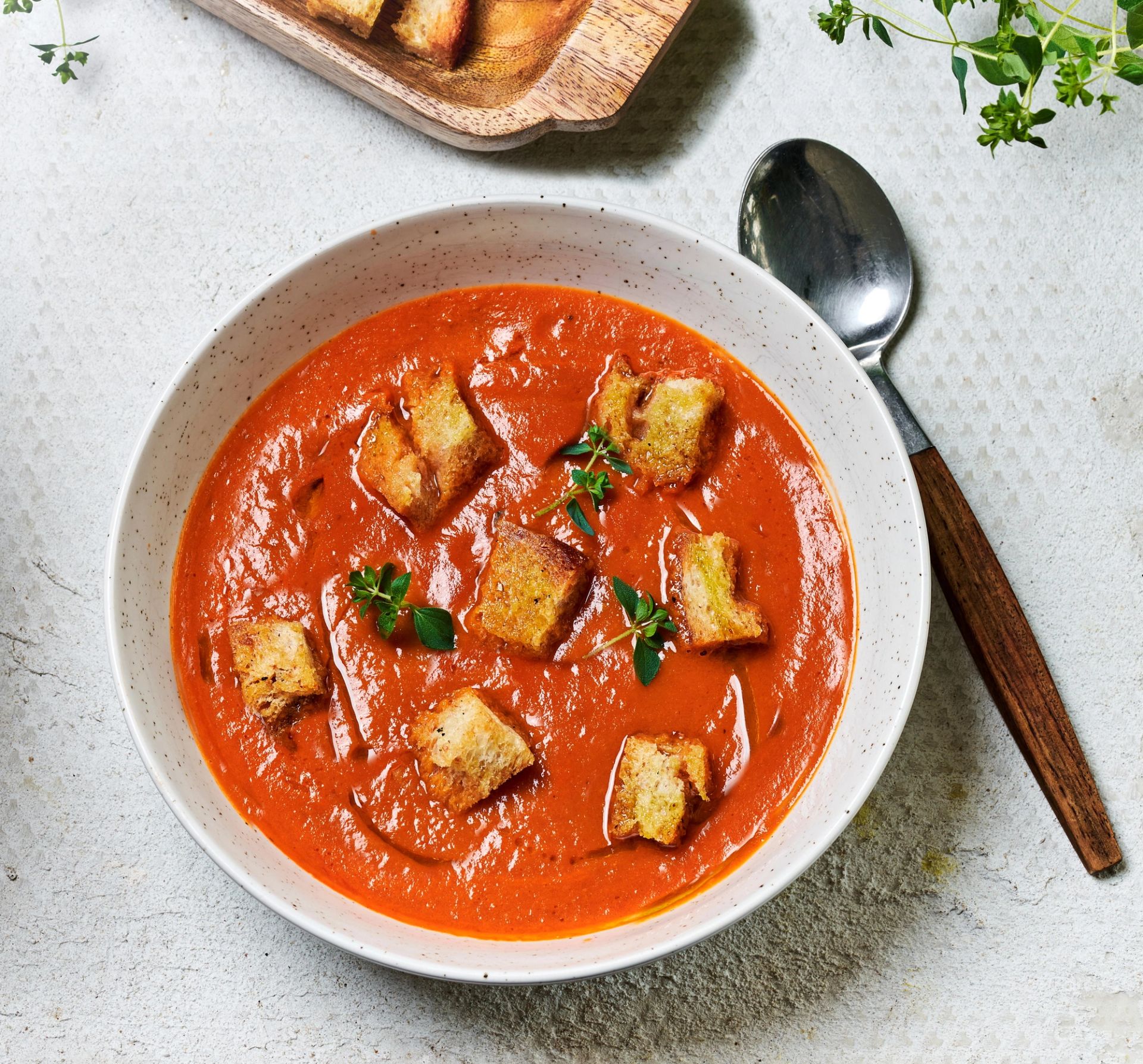 En skål med kremet tomatsuppe toppet med krutonger og friske urter, med en skje og ekstra krutonger ved siden av på en lys overflate.
