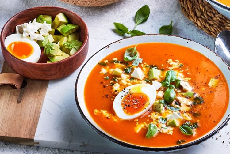 Paprika- og tomatsuppe i en skål, toppet med avokadobiter, smuldret fetaost, basilikum og et halvt kokt egg med rennende eggeplomme, servert med en liten skål avokado, egg og feta på siden.