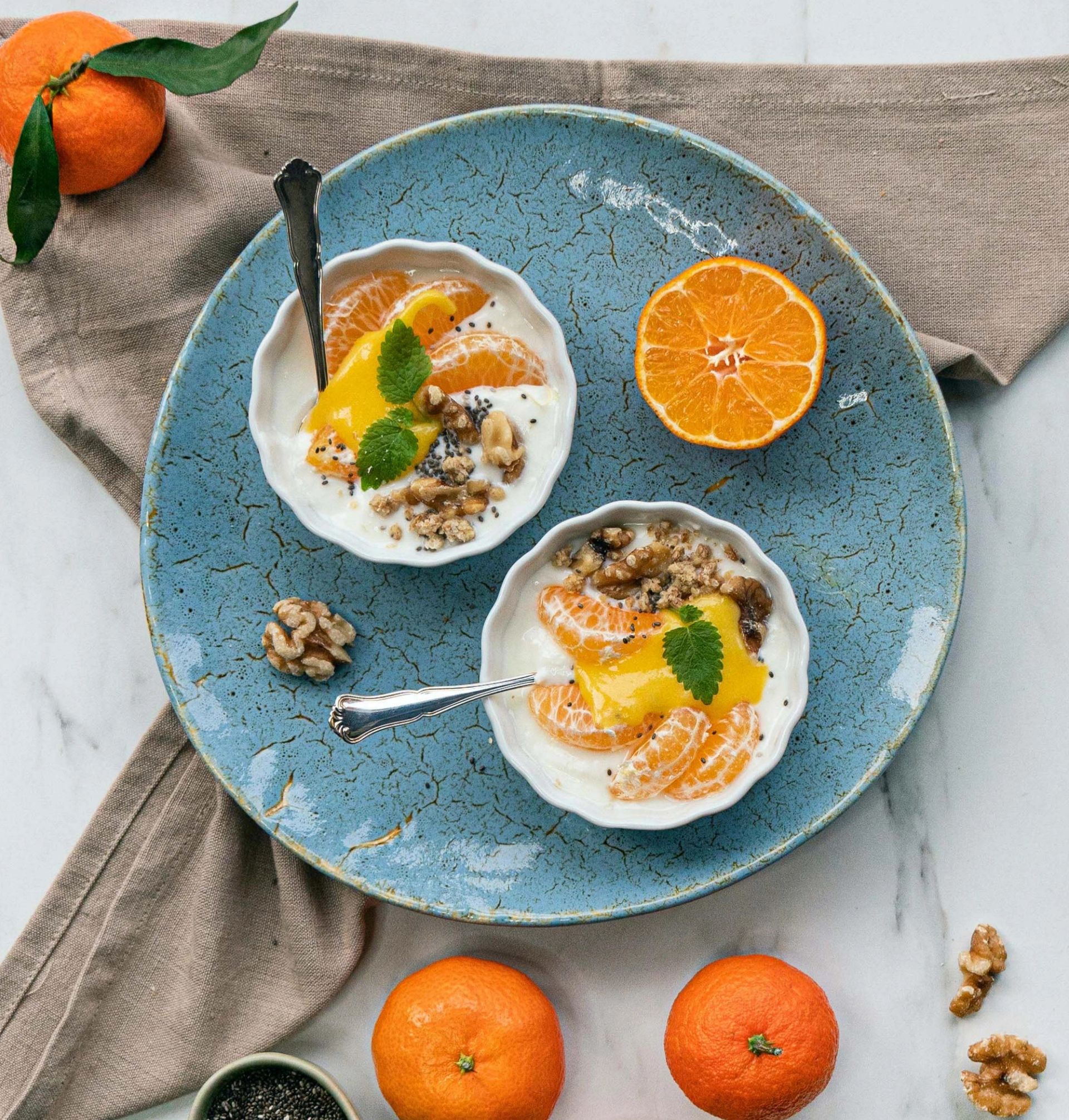 To små skåler med yoghurt toppet med mandariner, granola, nøtter og frisk mynte står på et stort, blått fat sammen med en halvert mandarin; rundt fatet ligger hele mandariner og valnøtter på en lys bordflate.