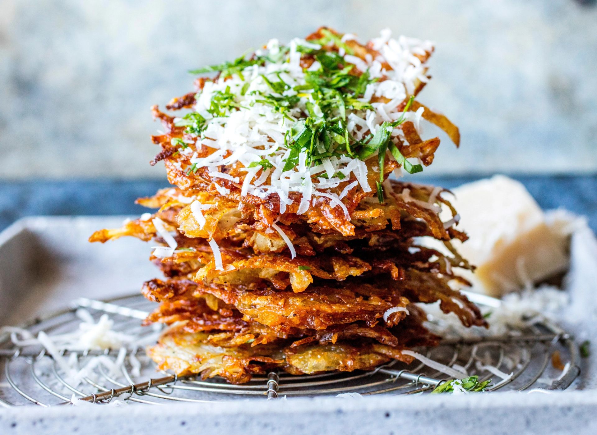 En høy stabel med gylne, sprøstekte røstipoteter som ligger på en bakerist. Stabelen er toppet med rikelig med revet parmesan og finhakkede grønne urter.