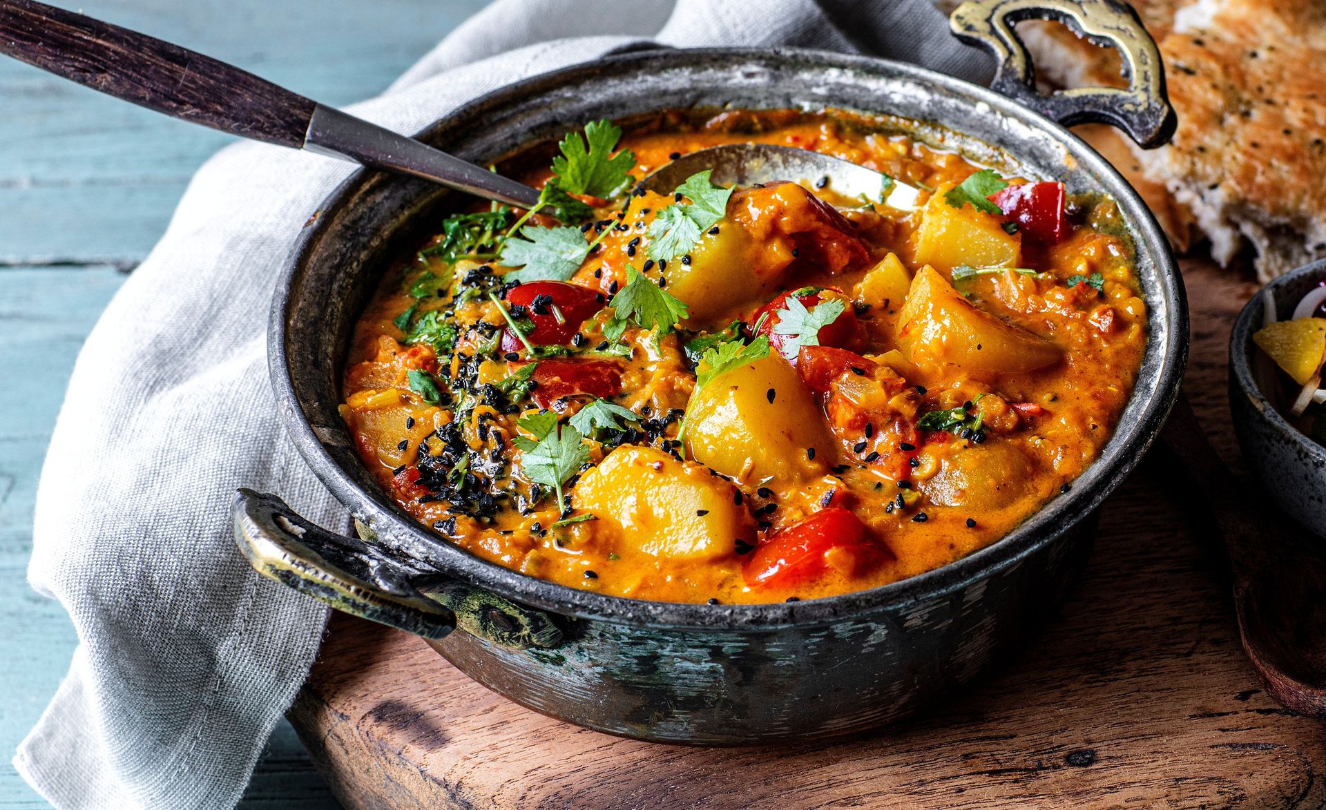 Fyldig gryte med kremet, oransje saus servert i en mørk jerngryte, fylt med store biter av potet, paprika og linser, toppet med frisk koriander og svarte frø, satt på et rustikt trebord med en skje i gryten.