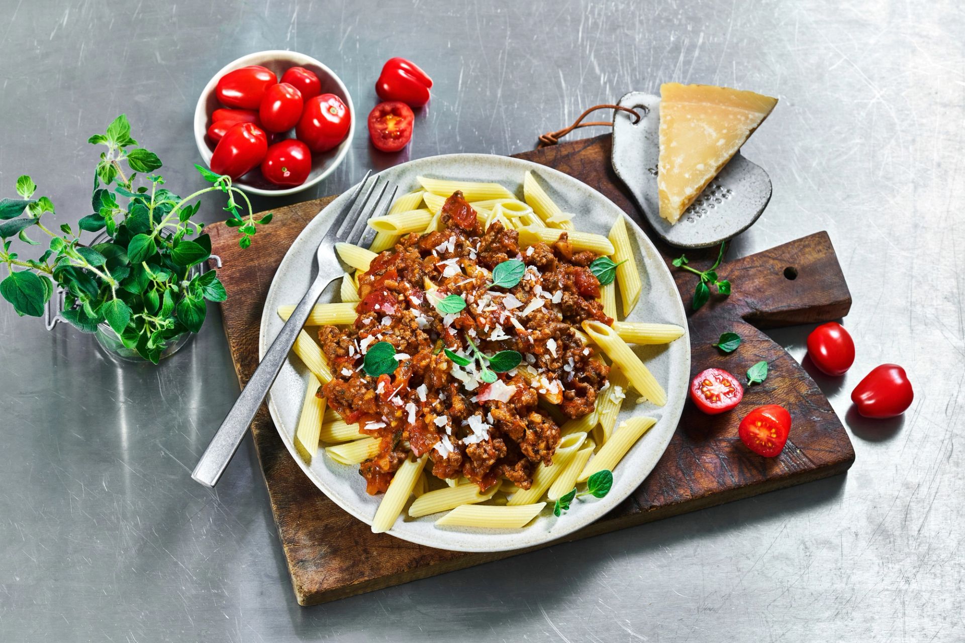 En tallerken med pennepasta toppet med kjøtt- og tomatsaus, revet parmesan og friske urter, servert på et trefjøl med cherrytomater, parmesanost og urter rundt.