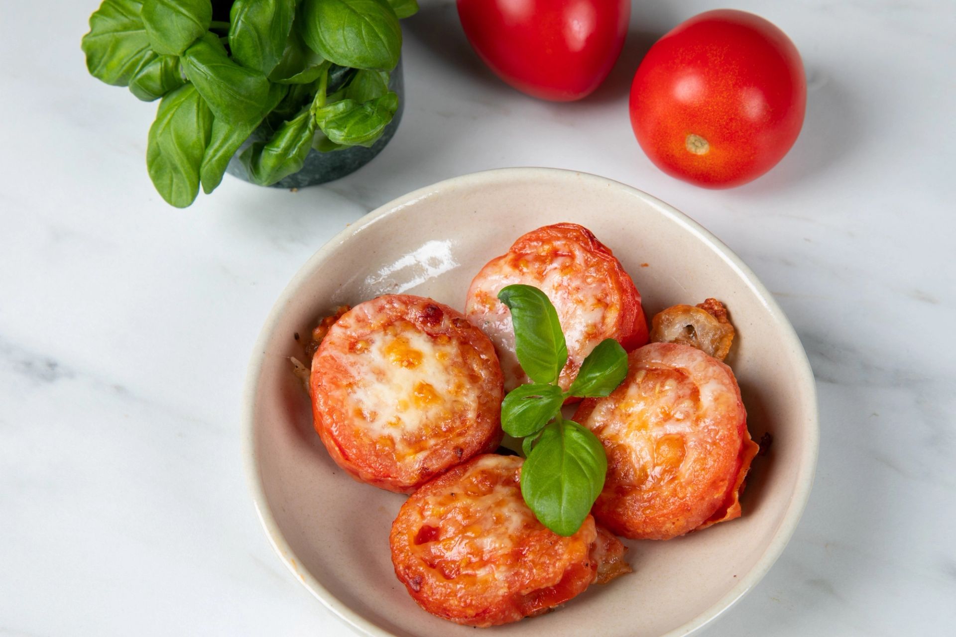 Gratinerte tomatskiver toppet med smeltet ost og frisk basilikum, servert på en lys tallerken. Friske tomater og en potte basilikum ligger i bakgrunnen på en hvit benkeplate. Enkel og sunn airfryer‑oppskrift.