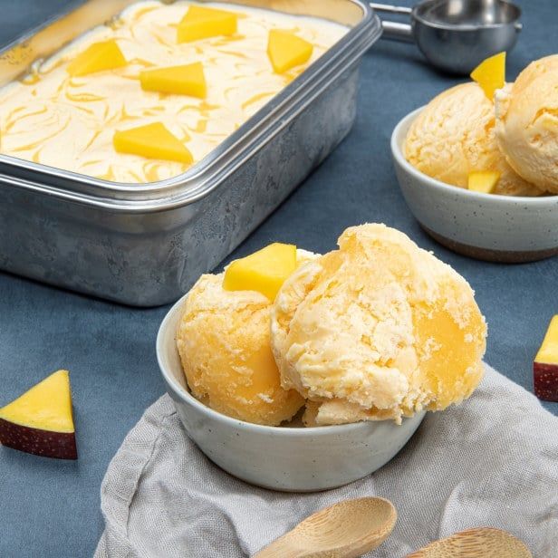 Kremet mangoiskrem servert i skål, toppet med ferske biter av mango ved siden av iskrembeholder.