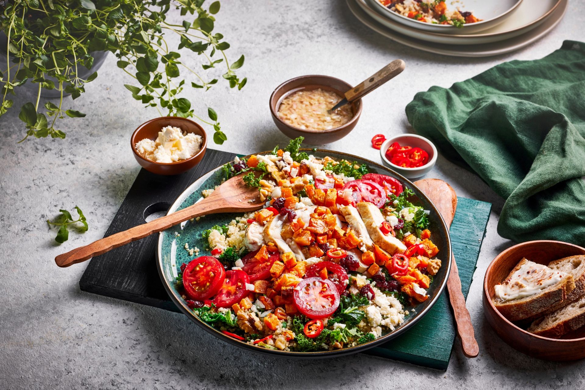 Stor bolle med fargerik salat av grønne bladgrønnsaker, korn, bakte søtpoteter, cherrytomater og skiver av grillet kylling, toppet med friske urter og chili. Servert med brød og dressing i skåler rundt på lys bakgrunn.s