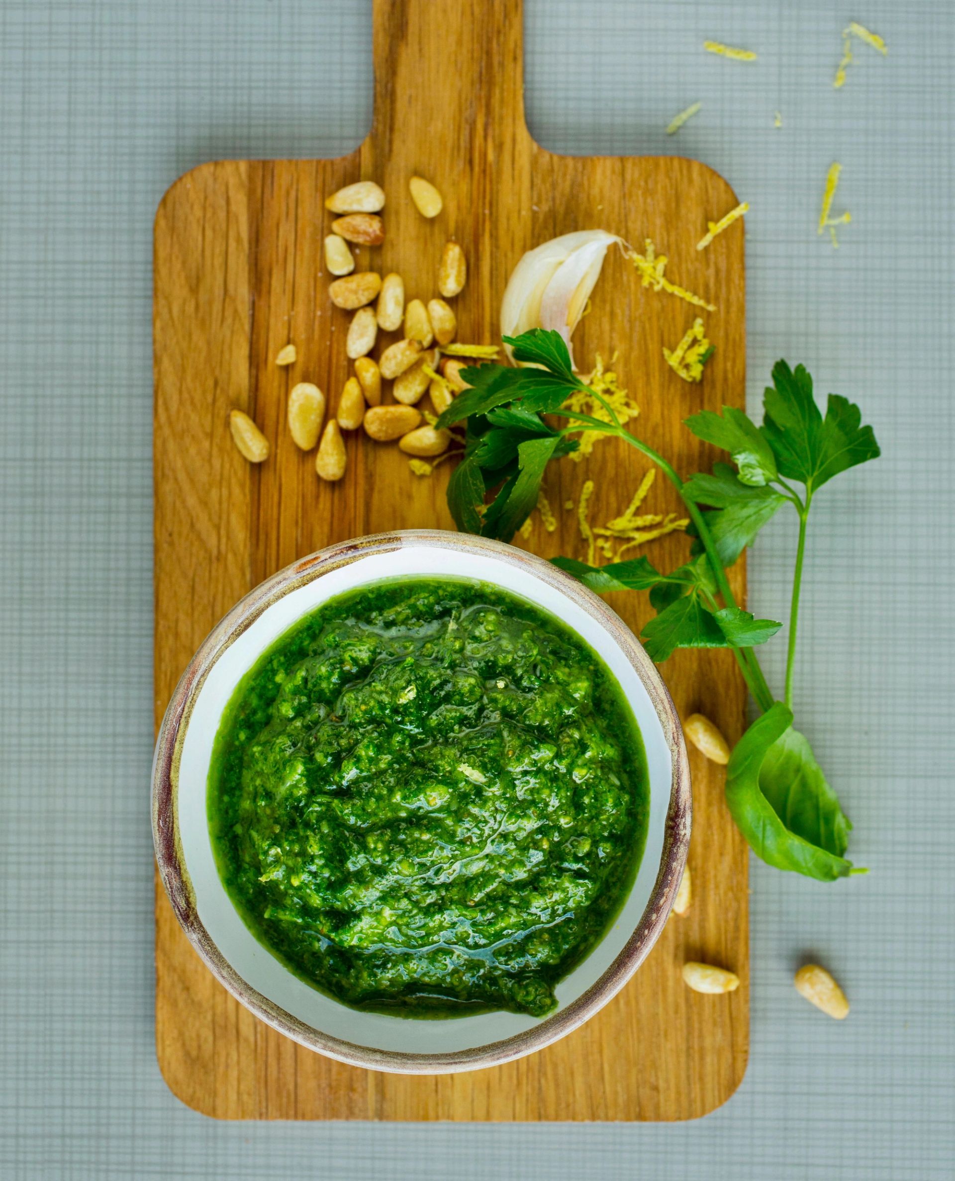 En bolle med grønn persillepesto står på et lite skjærebrett av tre. Rundt bollen ligger hele mandler, hvitløksfedd, sitronskallstrimler og noen kvister fersk persille.