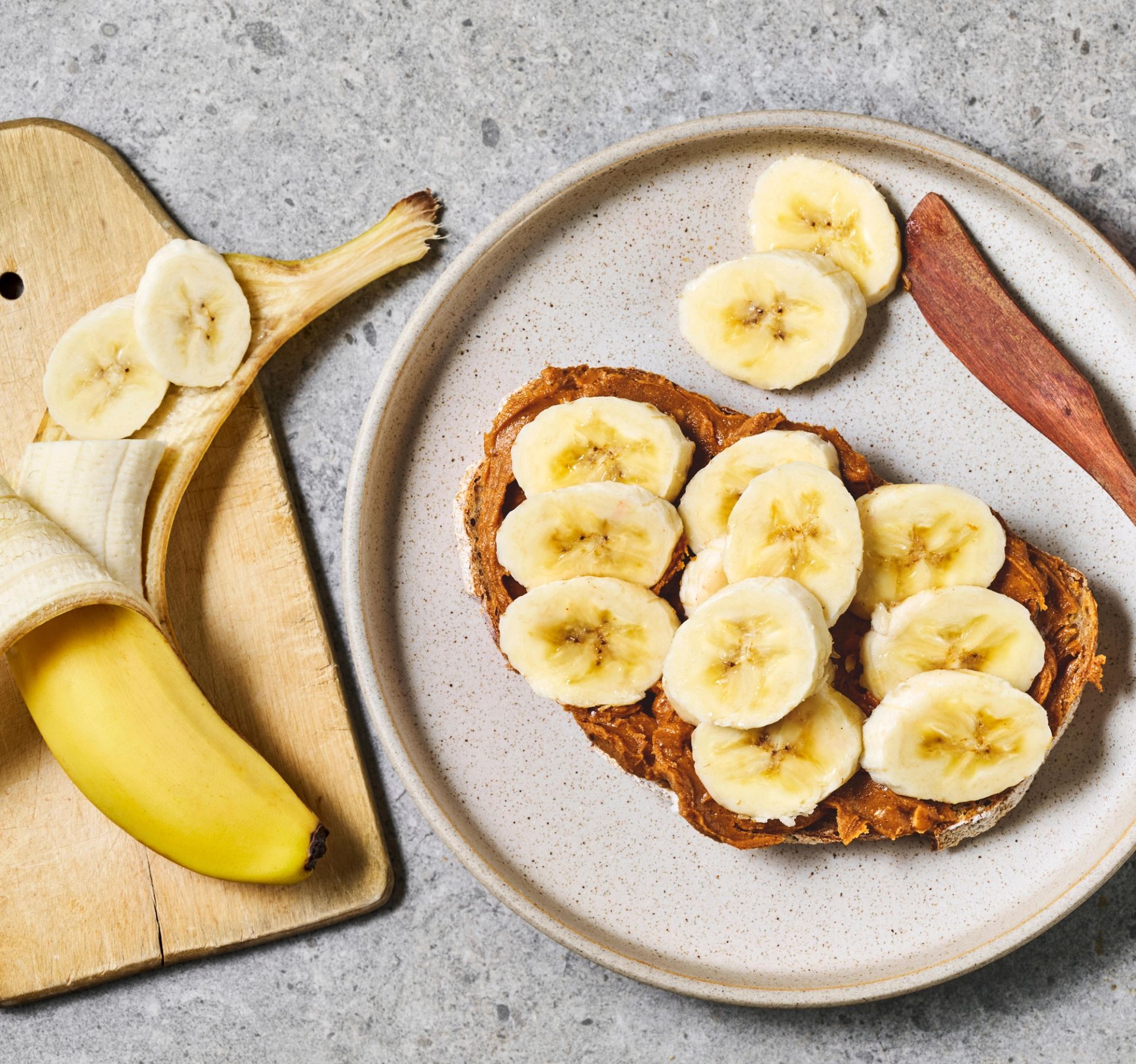 Brødskive med nøttepålegg og bananskiver er servert på en tallerken som et enkelt mellommåltid eller frokost. Ved siden av ligger en halvskrelt banan på et skjærebrett og noen ekstra bananskiver, klare til å legges på brødskiven.