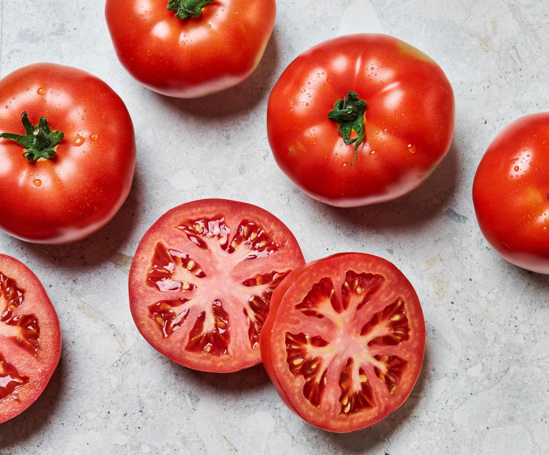 Flere hele bifftomater og to skiver av en oppskåret bifftomat ligger på en lys steinoverflate. De hele tomatene er runde og røde med grønne stilker, mens de kuttede skivene viser den saftige, mønstrede innmaten.