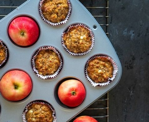 Grove eplemuffins i papirformer i en muffinsform på et bakerist, med friske røde epler rundt, avkjølt på et mørkt bord.