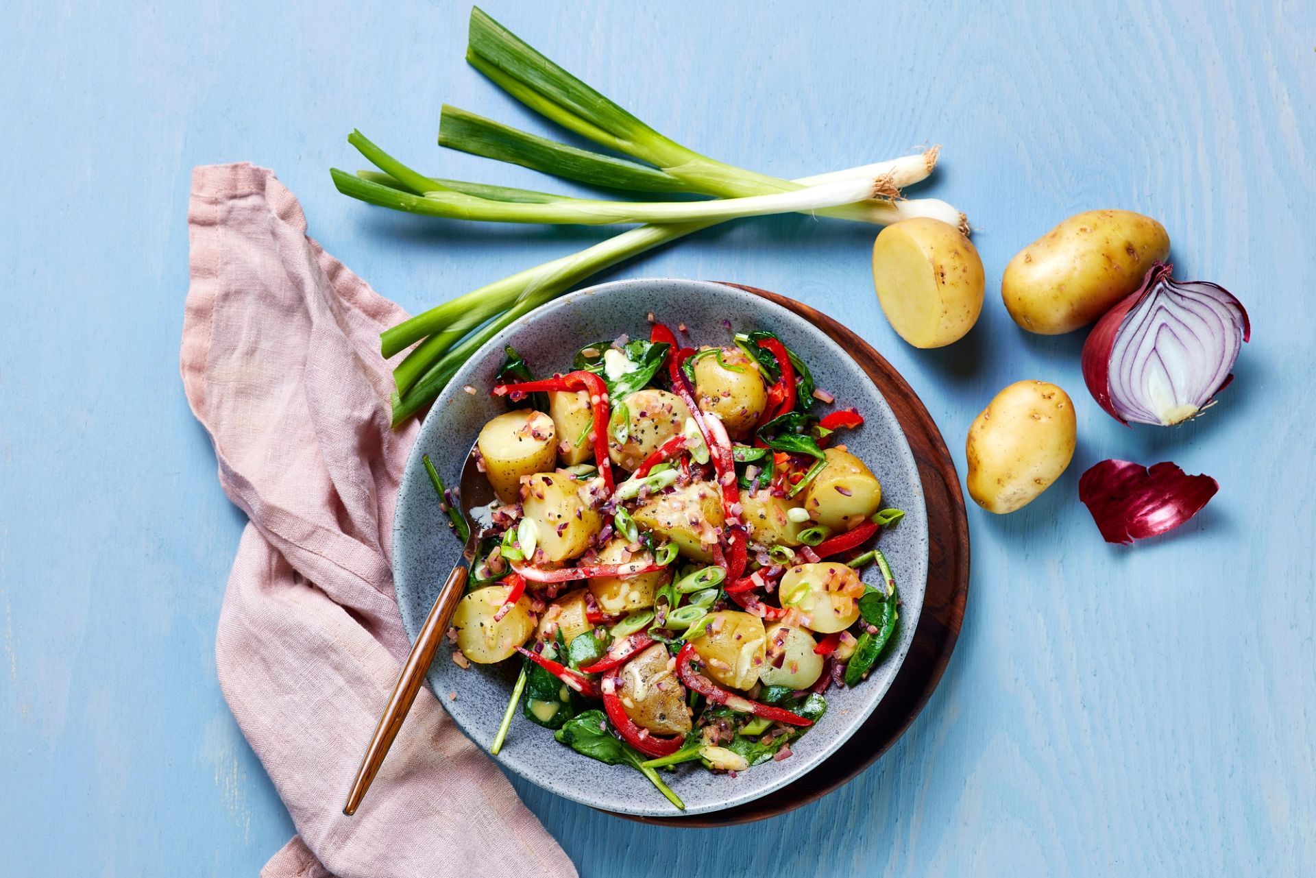 En bolle med potetsalat med poteter, paprika, rødløk og grønt, omgitt av rå ingredienser som vårløk, poteter og halv rødløk på et lyseblått bord.