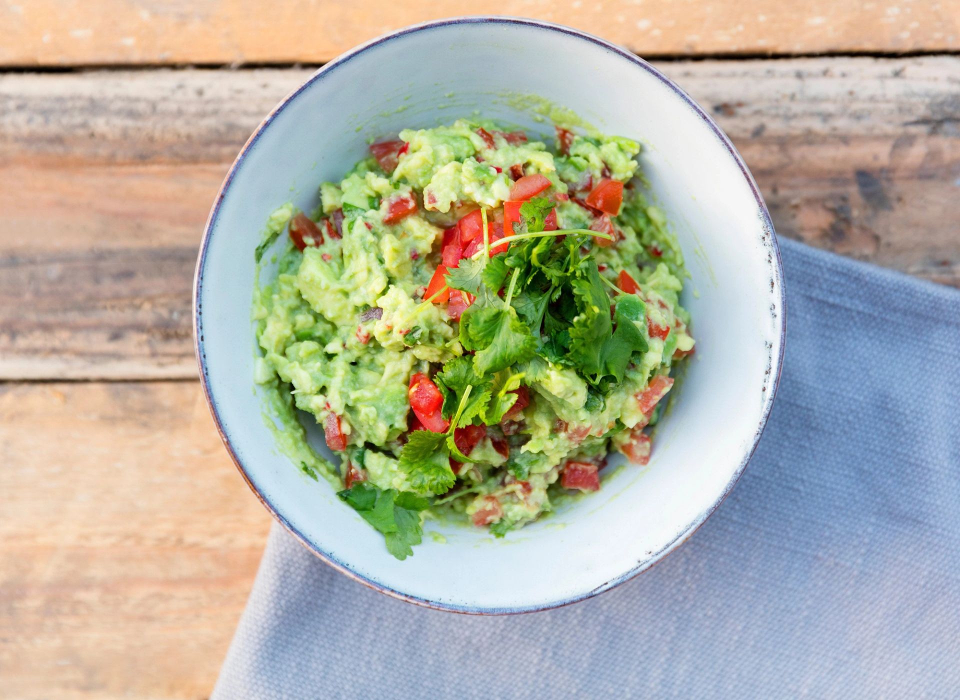 Bolle med guacamole laget av avokado, lime, plommetomat og koriander, servert på et trebord med en grå duk under.