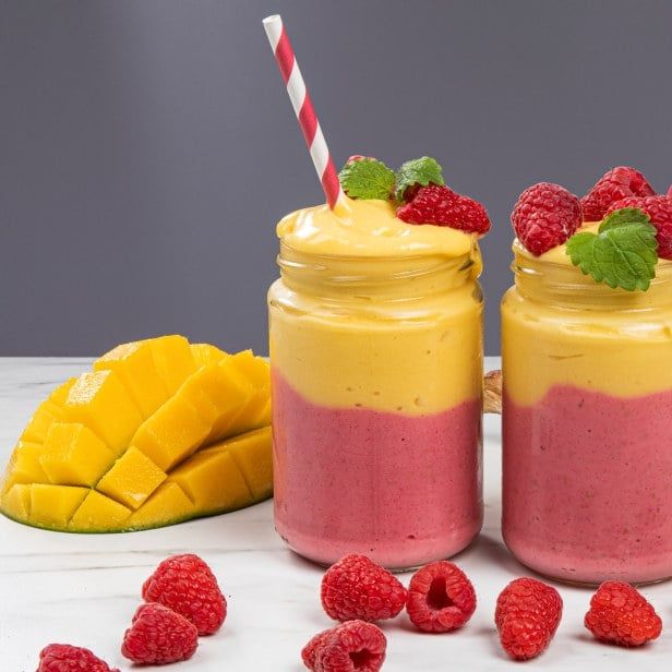 Sunrise smoothie i lag med gul mango og rosa bringebær, servert i glass med sugerør, toppet med friske bringebær og mynte, med mango og bringebær ved siden av.