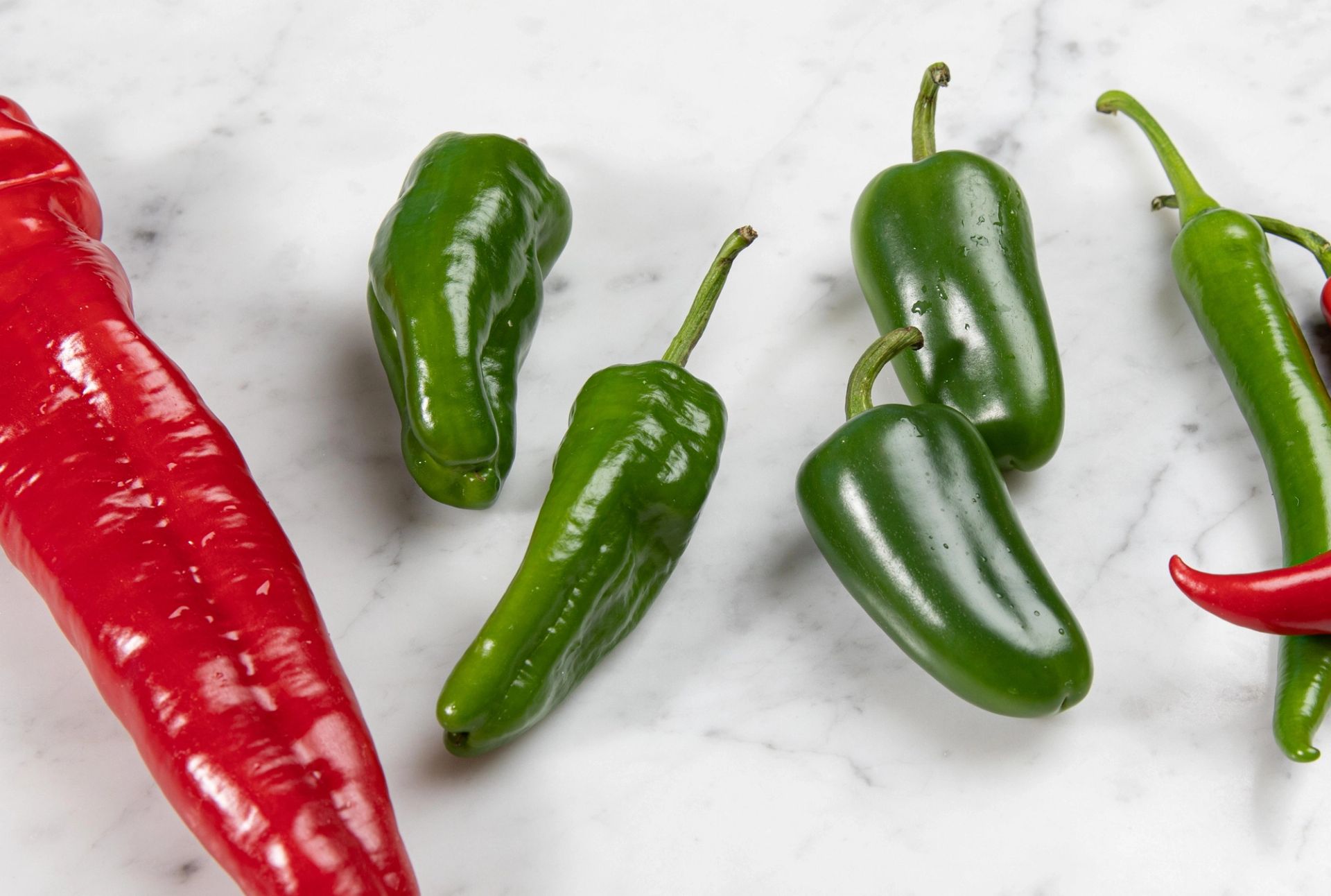 Flere grønne jalapeñoer og en stor rød chili ligger spredt på en lys marmorlignende overflate.