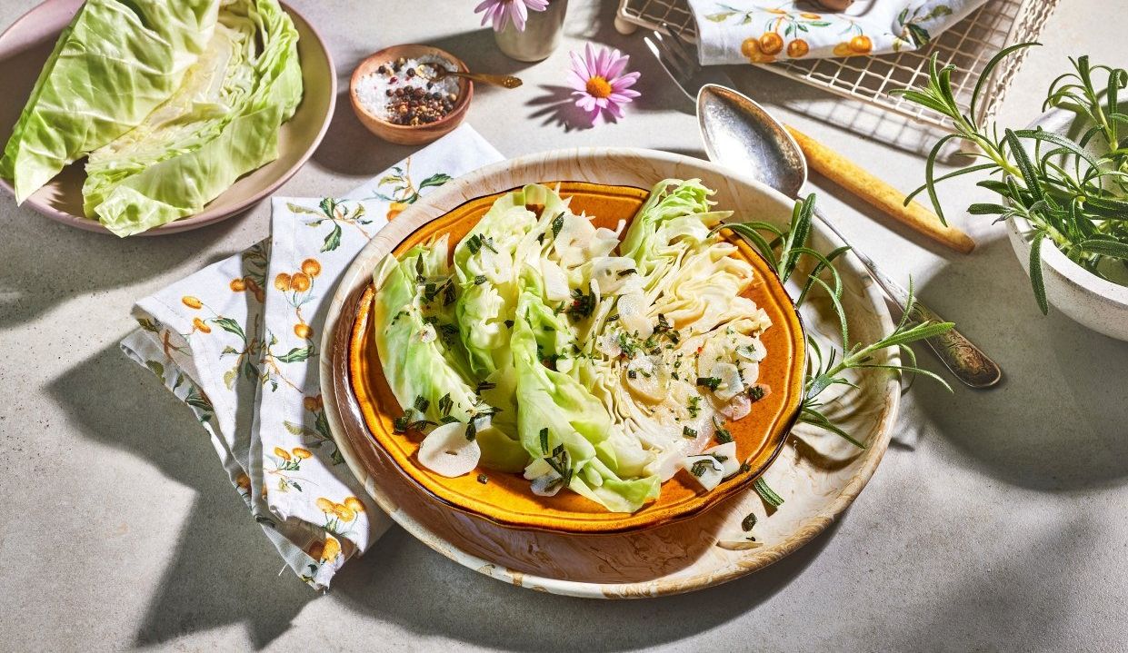 Smørdampet spisskål servert på et fat, toppet med skiver av hvitløk og frisk rosmarin, omgitt av rå spisskål, krydder og urter på et lyst kjøkkenbord.