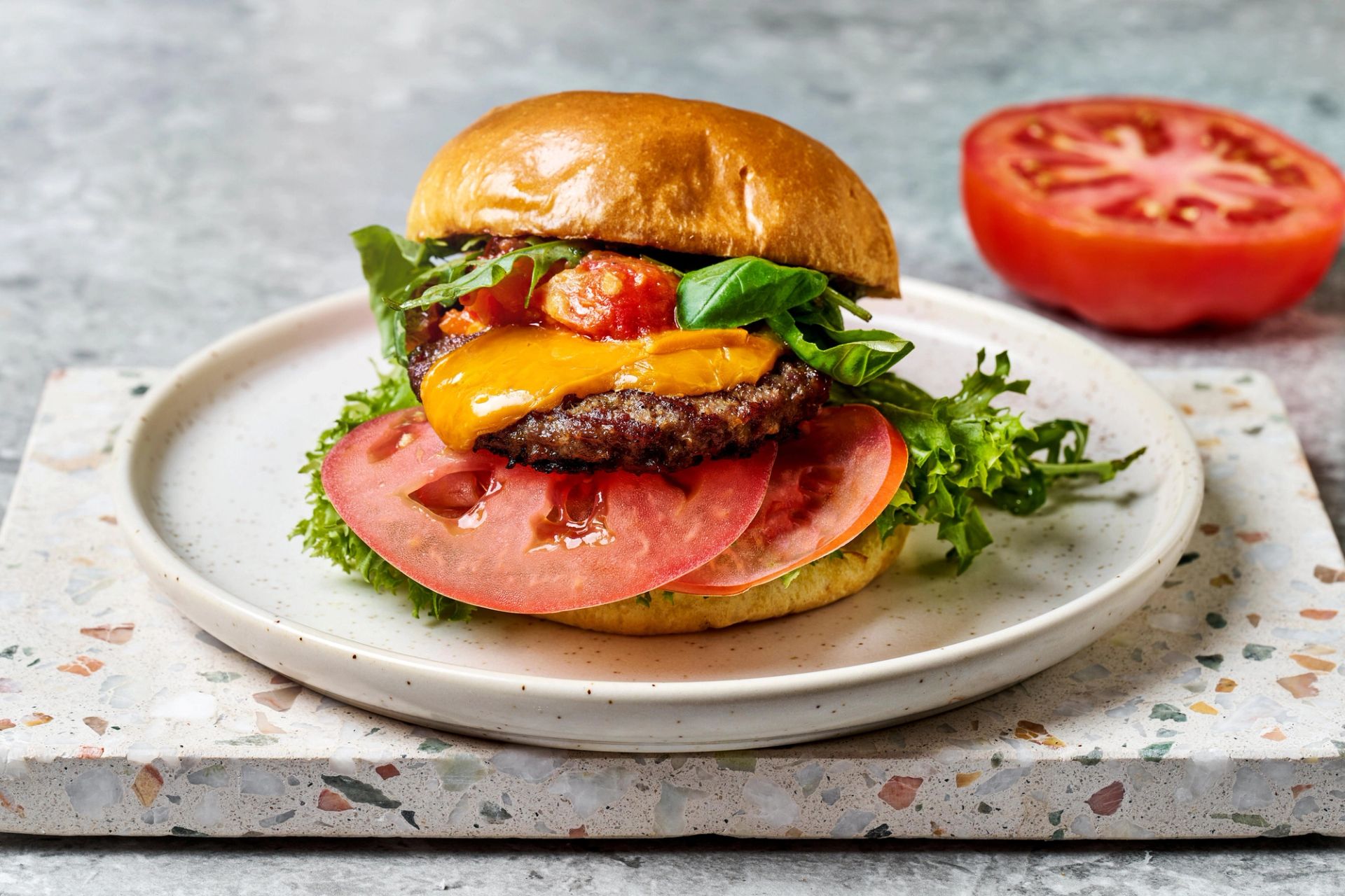 Hamburger med saftig kjøttburger, smeltet cheddarost, tykke skiver tomat, salat og tomatrelish i et glinsende burgerbrød. Burgeren er servert på en lys tallerken med grønn salat ved siden av, og en halv tomat ligger i bakgrunnen.