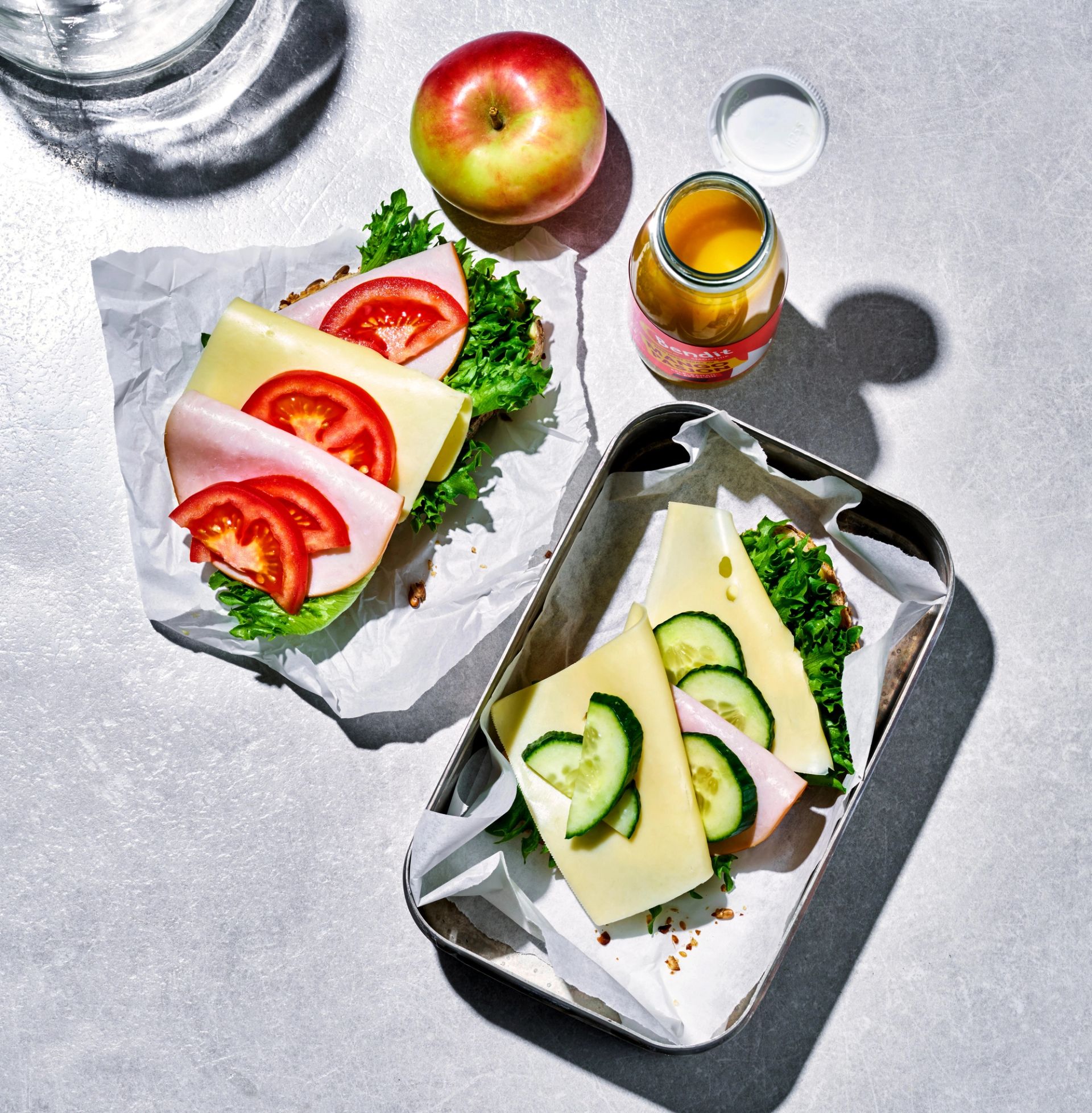 To grove brødskiver med ost, skinke, salat, tomat og agurk er pakket inn som matpakker og servert sammen med et eple, et glass juice og et glass vann. Måltidet er arrangert på et lyst underlag og gir et enkelt og næringsrikt inntrykk av lunsj eller skolemat.