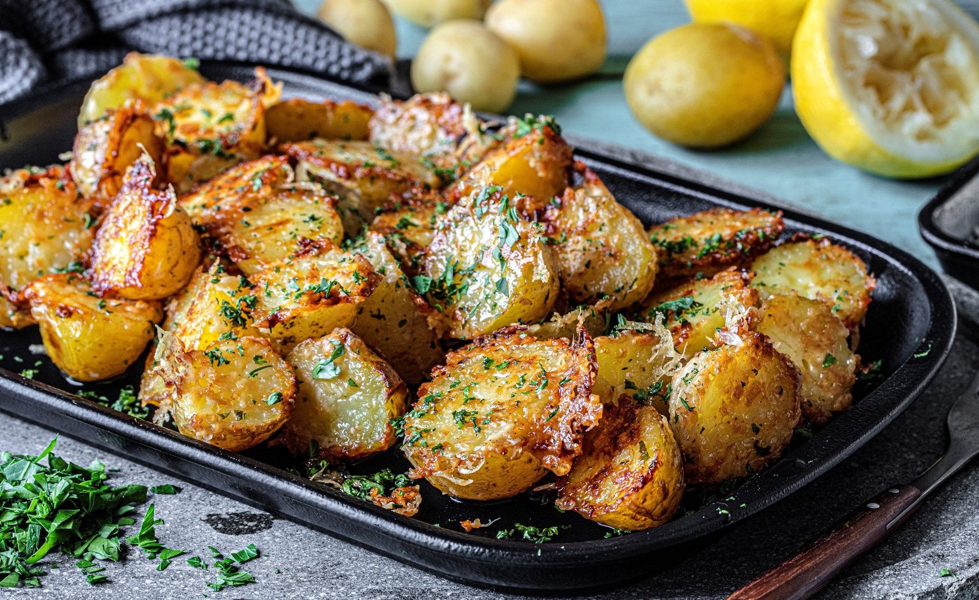 Stekte parmesanpoteter i en svart langpanne, med gylne, sprø potethalvdeler dekket av smeltet parmesan og finhakkede urter. Rundt formen ligger hele poteter og en halv sitron på et rustikt kjøkkenbord.
