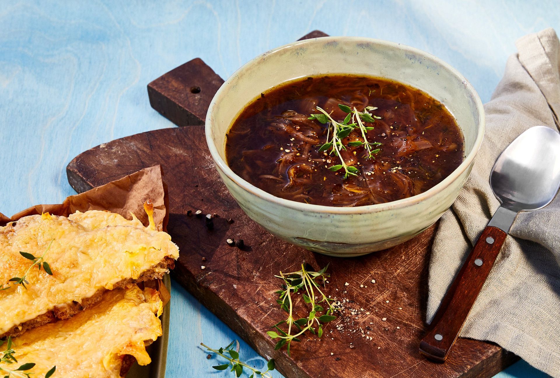 Fransk løksuppe med karamellisert løk i kraft toppet med urter, servert med gratinert ostesmørbrød ved siden av.