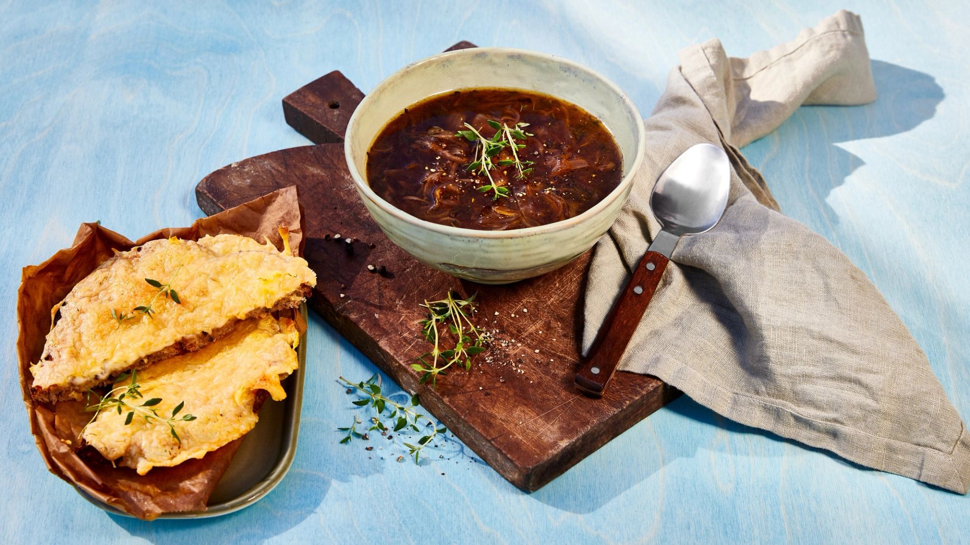 Fransk løksuppe med karamellisert løk i kraft toppet med urter, servert med gratinert ostesmørbrød ved siden av.