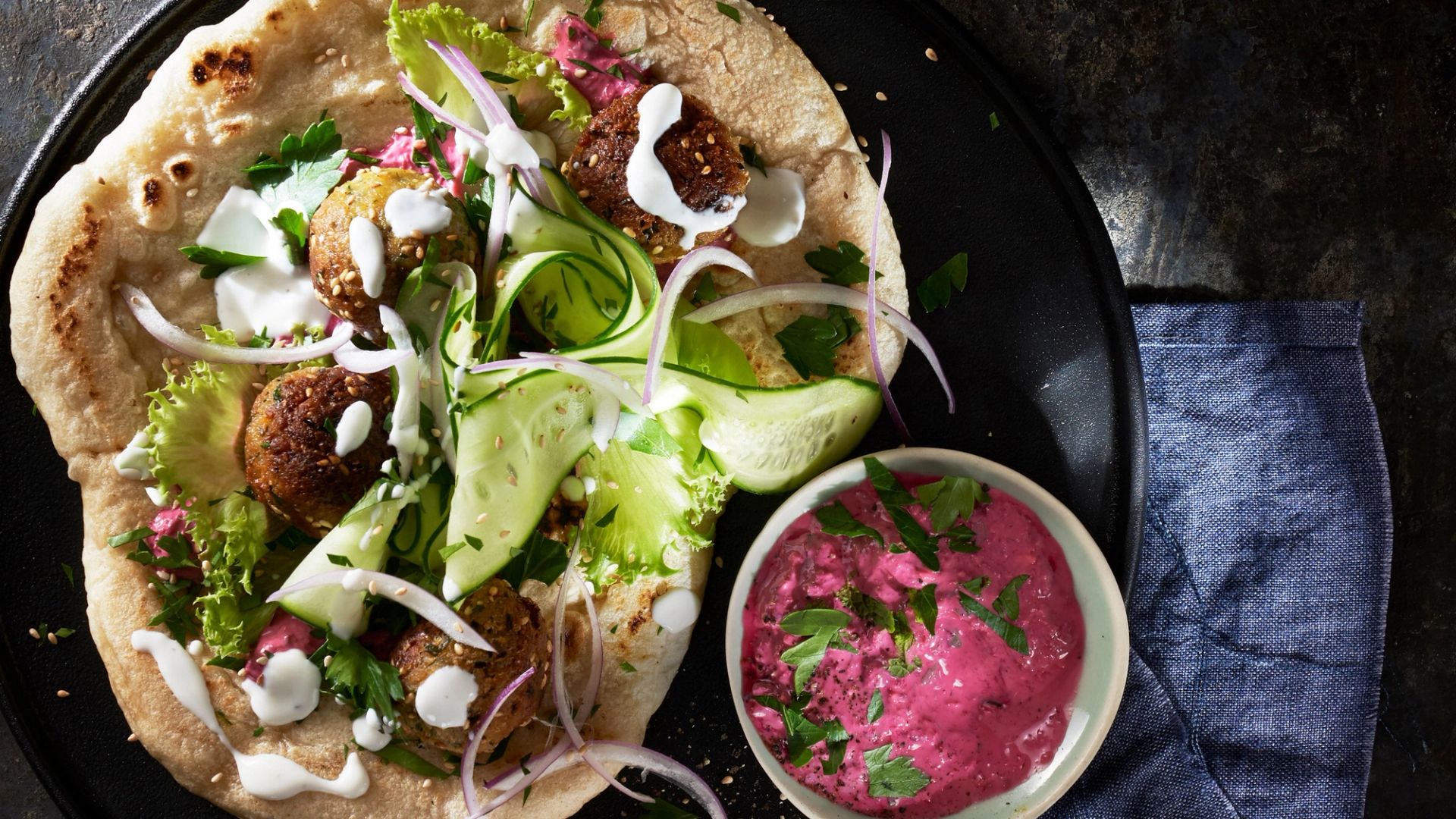 Et mørkt serveringsfat med et pitabrød toppet med falafel, grønne blader, agurkskiver, rødløk og hvit saus, servert ved siden av en liten skål med rødbet-tzatziki med persille.