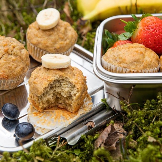 Grove muffins med bananskiver på toppen, ligende på et brett i lyngen. Rundt muffinsene ligger blåbær og jordbær.
