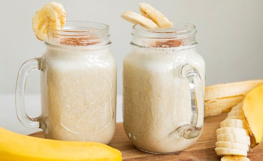 To glasskrukker fylt med kremet banansmoothie står på en trefjøl, pyntet med hele og oppskårne bananer.