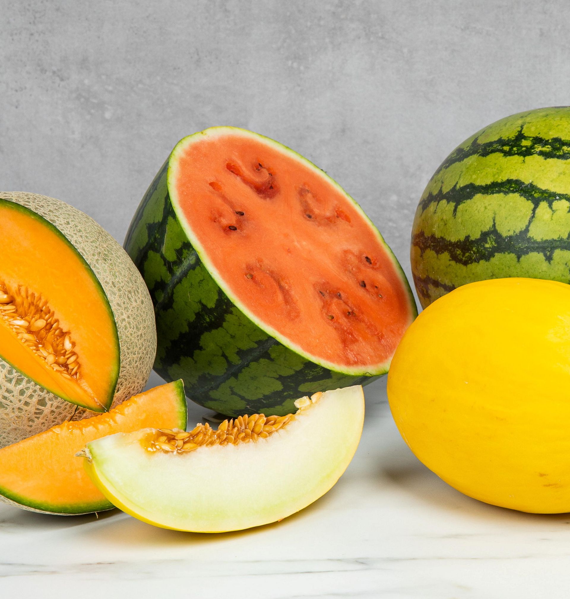 Ulike meloner arrangert på en lys overflate, inkludert vannmelon hel og delt i to, cantaloupe med oransje fruktkjøtt og frø, samt gul melon og lyse melonbåter mot en nøytral bakgrunn.