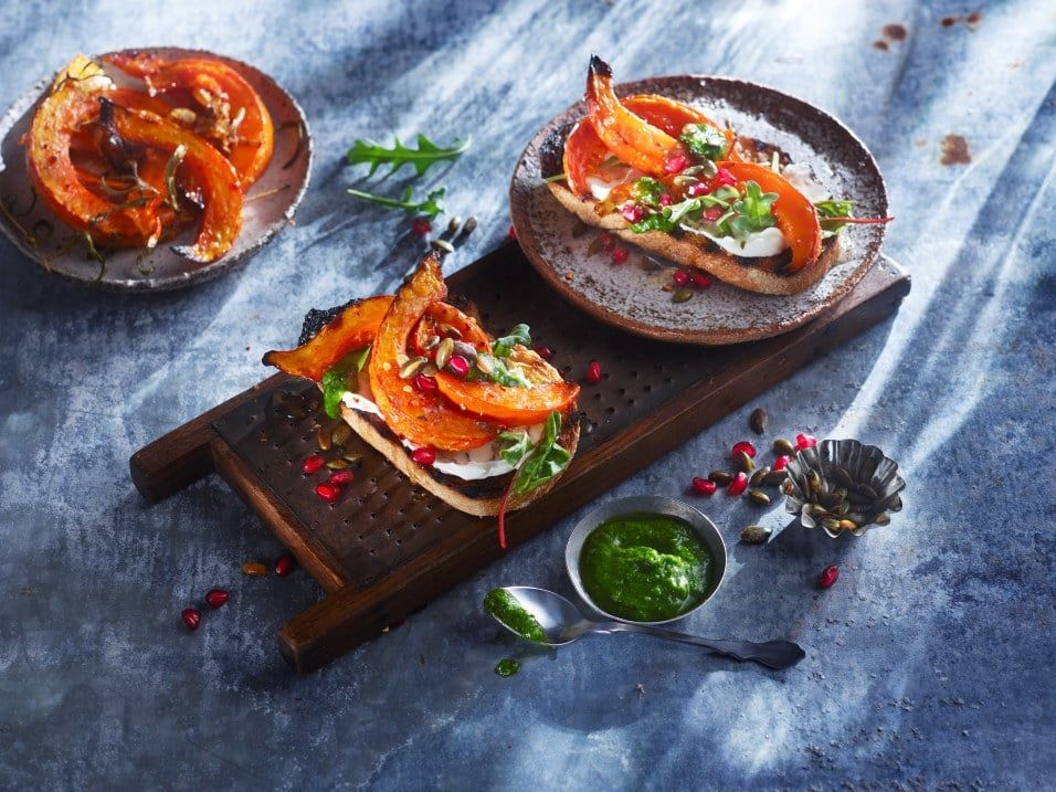 To sprø bruschettaer toppet med ovnsbakte gresskarskiver, kremet ost, granateplekjerner og gresskarfrø, anrettet på en mørk trefjøl. Ved siden av står en liten skål med grønn pesto.