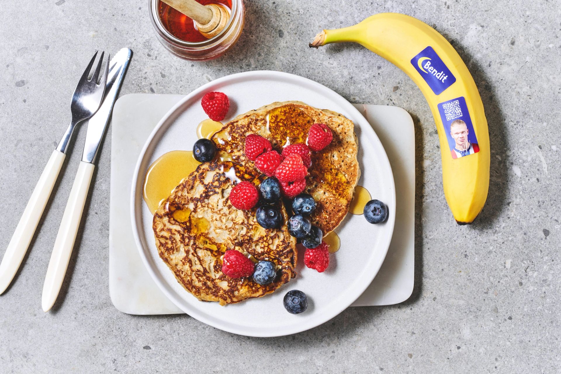 Stabel med havre- og bananpannekaker toppet med bananskiver, bringebær og blåbær, servert med sirup, friske bær og bananer på et bord.