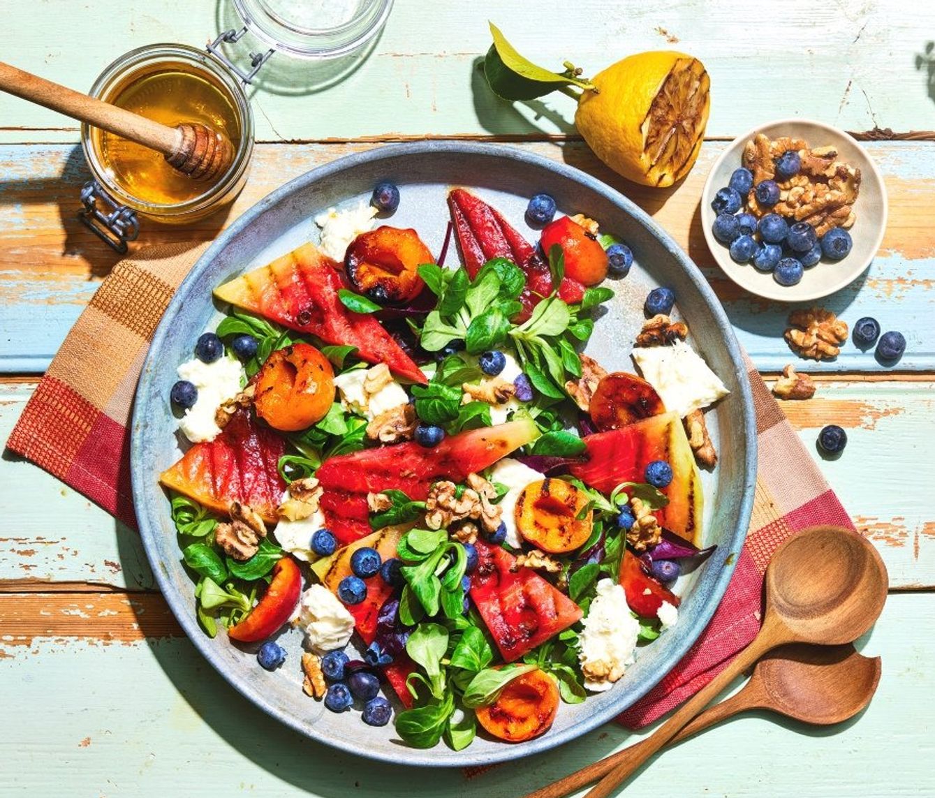 Fargerik salat servert på et stort fat med biter av vannmelon, grillede nektariner, blåbær, valnøtter, grønne salatblader og smuldret hvit ost. Rundt fatet står små skåler med blåbær og nøtter, en halv sitron og et glass med honning på et lyst, rustikt trebord. Den friske kombinasjonen av frukt, bær og grønnsaker gir retten et sommerlig og innbydende uttrykk.Gi tilbakemelding om BizChat.