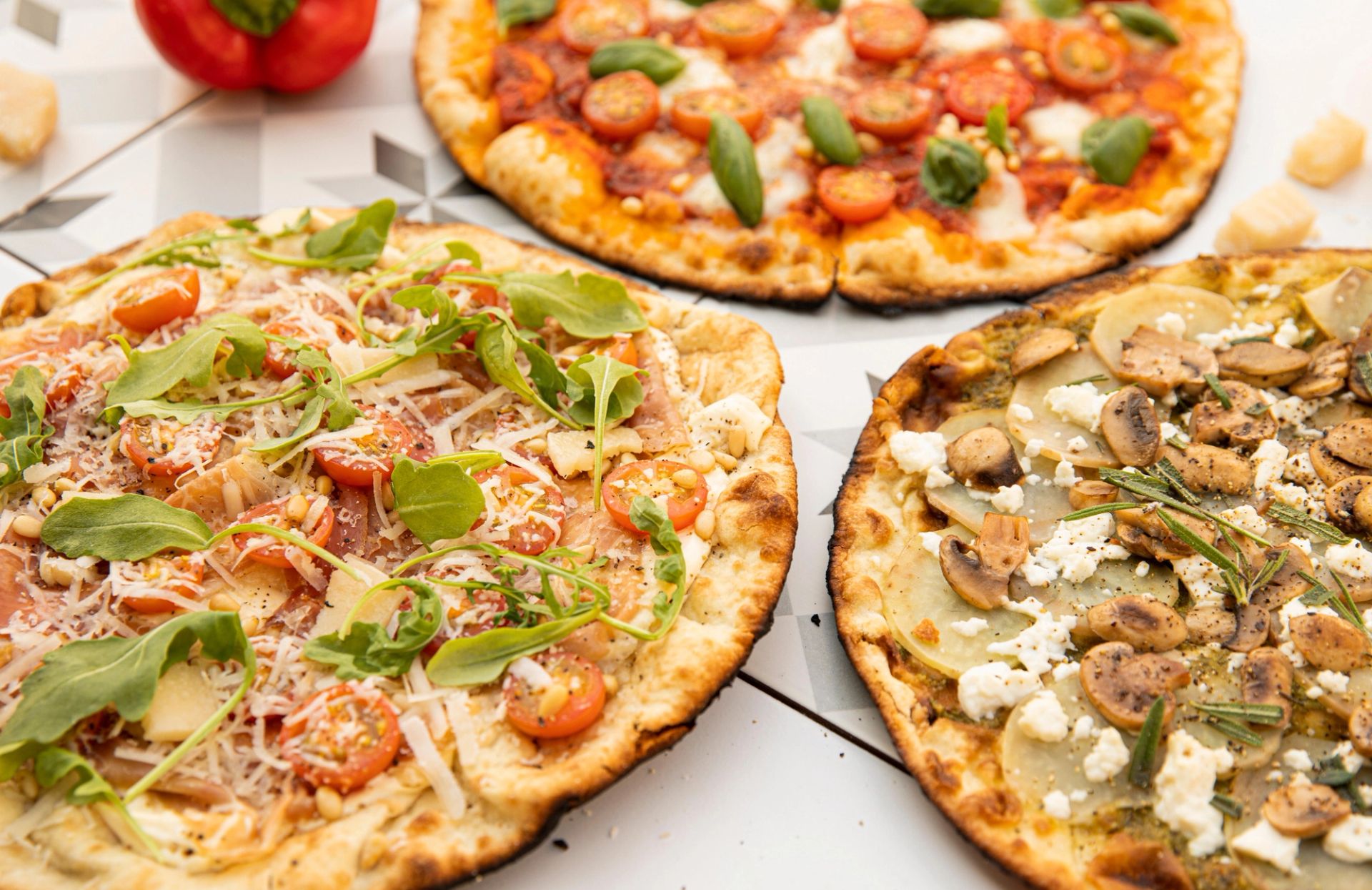 Tre ulike pizzaer med variert topping som ruccola, cherrytomater, ost, sopp, potet og urter, servert på et bord med spredte grønnsaker og ostebiter rundt.