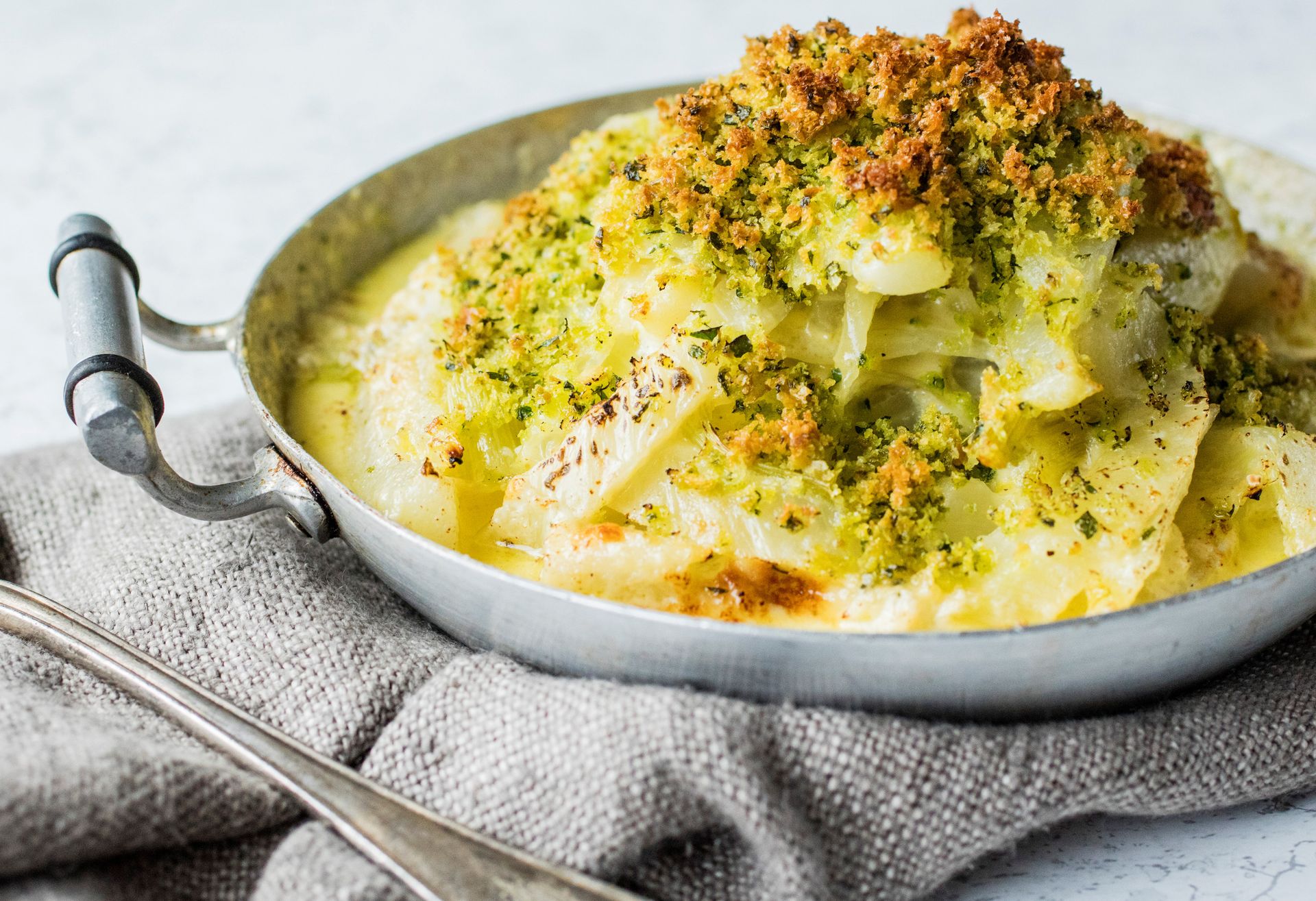 En liten, rund metallform fylt med gratinert fennikel i kremet saus, toppet med sprø, grønnkrydret brødsmuleblanding. Formen står på et grått, teksturert kjøkkenhåndkle, med en stor skje ved siden av.