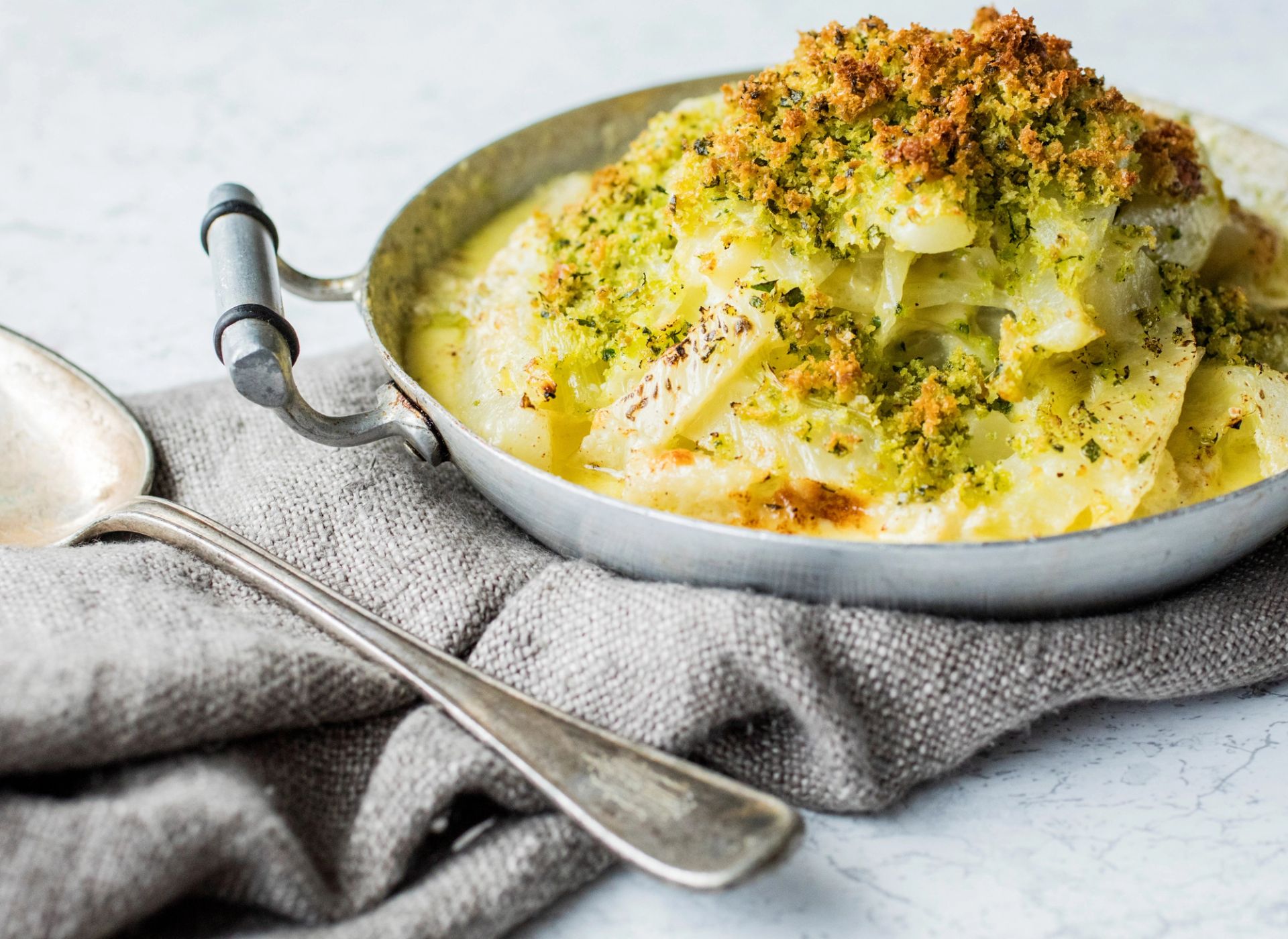 En liten, rund metallform fylt med gratinert fennikel i kremet saus, toppet med sprø, grønnkrydret brødsmuleblanding. Formen står på et grått, teksturert kjøkkenhåndkle, med en stor skje ved siden av.