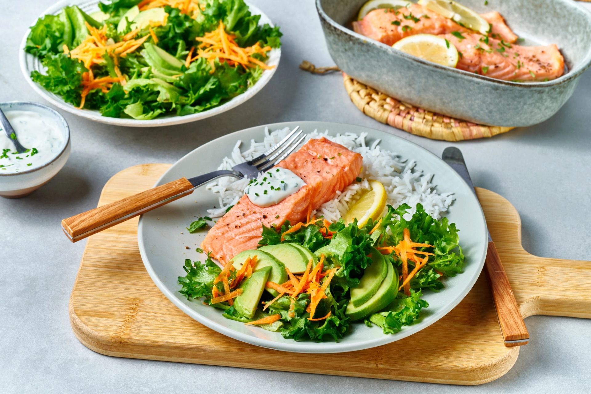 En ferdig anrettet middagstallerken med ovnsbakt laks servert på et trefjøl. Tallerkenen inneholder en rosa laksefilet toppet med en hvit urtedressing, hvit ris, en sitronbåt og en frisk grønn salat med avokadoskiver og revet gulrot.