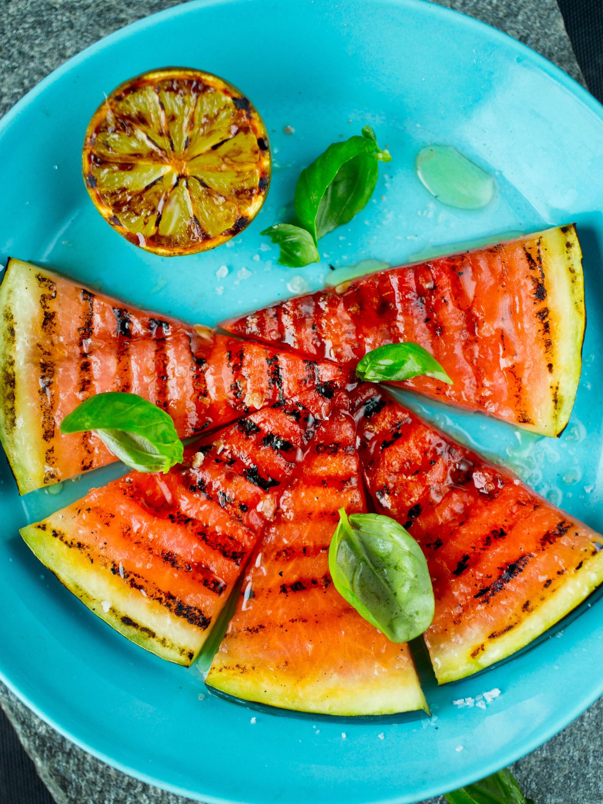Grillede vannmelonbåter arrangert i en sirkel på en turkis tallerken, med synlige grillstriper, dryss av salt og friske basilikumblader. En halvt grillet lime ligger øverst på tallerkenen.