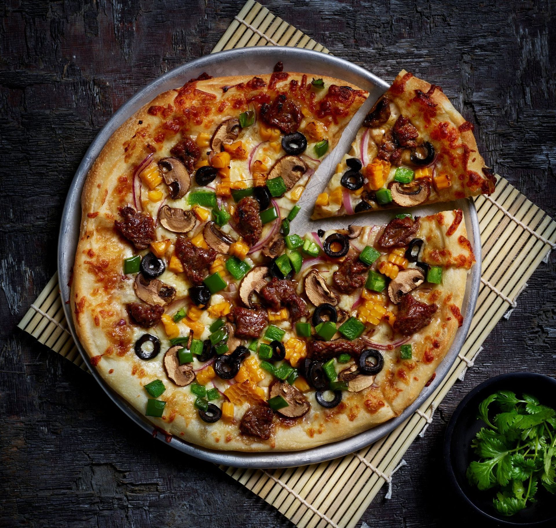 Nybakt pizza toppet med marinert oksekjøtt, sopp, rødløk, grønn paprika, mais og svarte oliven, servert på en metallplate over en bambusmatte. En bit er skåret ut, og en skål med koriander står ved siden av.