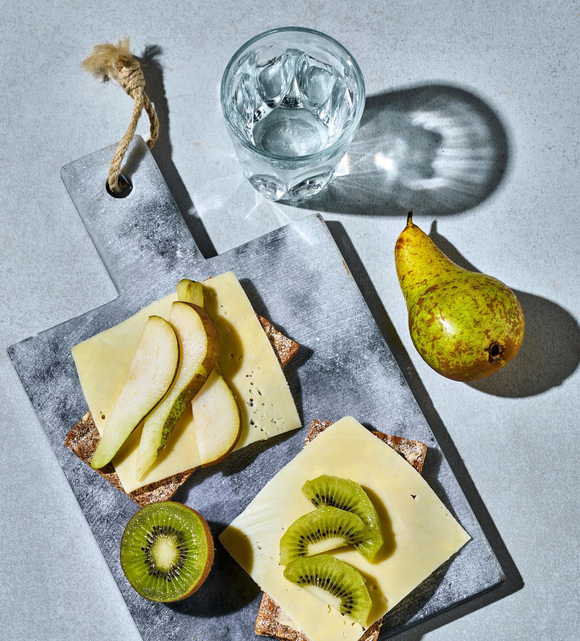 Et serveringsbrett med sprø knekkebrød toppet med ost og skiver av conference‑pære på det ene, og ost med kiwiskiver på det andre. Ved siden av står et glass vann som kaster en tydelig skygge, og en hel conference‑pære ligger på bordet.