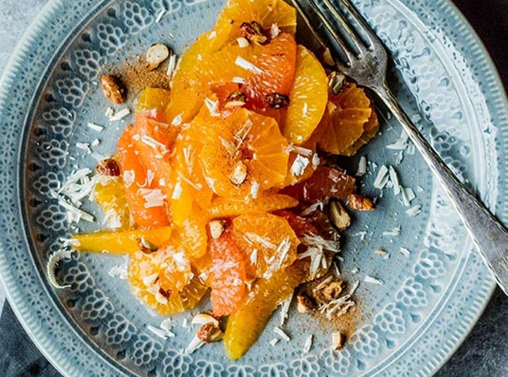 En frisk og fargerik sitrussalat servert på en dekorativ, blåmønstret tallerken. Salaten består av tynne skiver av appelsin og rød grapefrukt, toppet med hakkede nøtter, hvite sjokolade og et lett dryss av kanel.