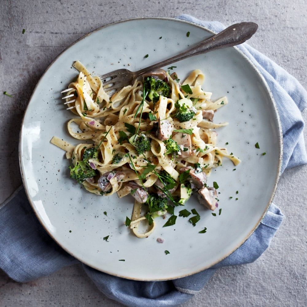 En tallerken med kremet pastarett laget av brede båndnudler, biter av sopp og broccoli, toppet med frisk urt. Tallerkenen står på en blå serviett på et lyst bord, med en gaffel i kanten.