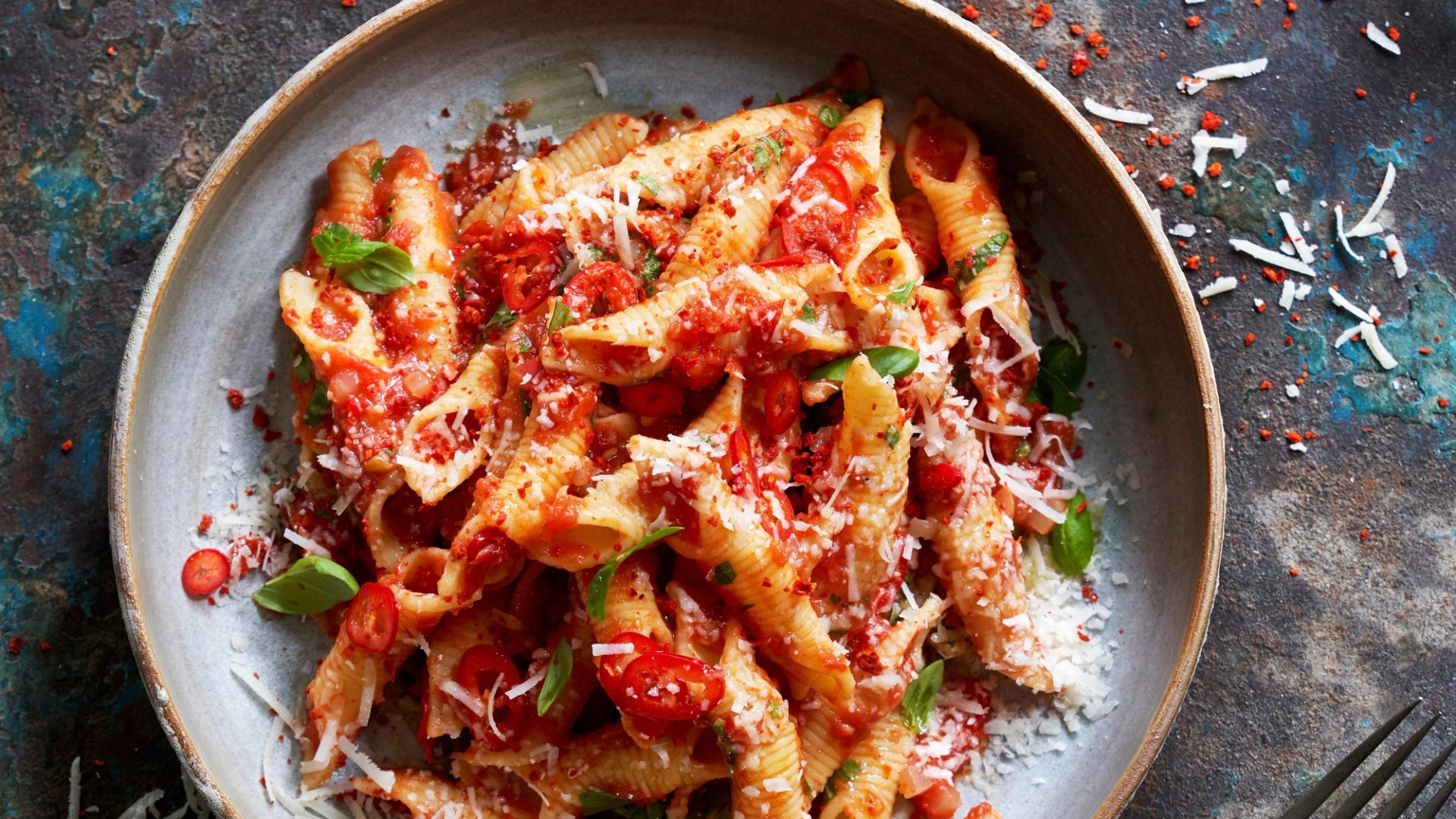 Skål med pasta (conchiglie) i tomatsaus, toppet med revet parmesan, friske basilikumblader og skiver av chili, fotografert ovenfra på mørk bakgrunn.